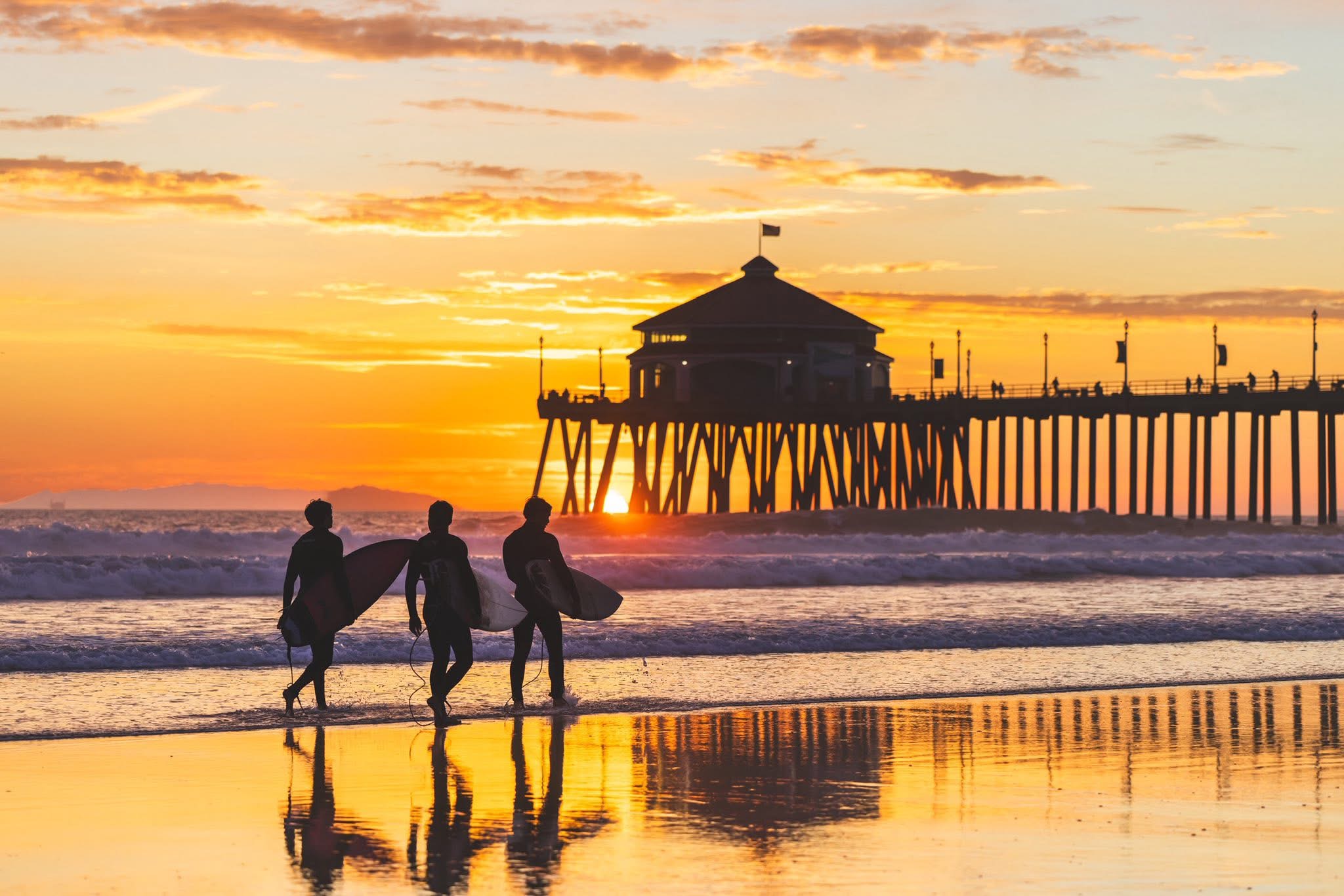 Surfers carrying boards at golden hour with Huntington Beach Pier in the background — 1 mile from 19621 Trident Ln